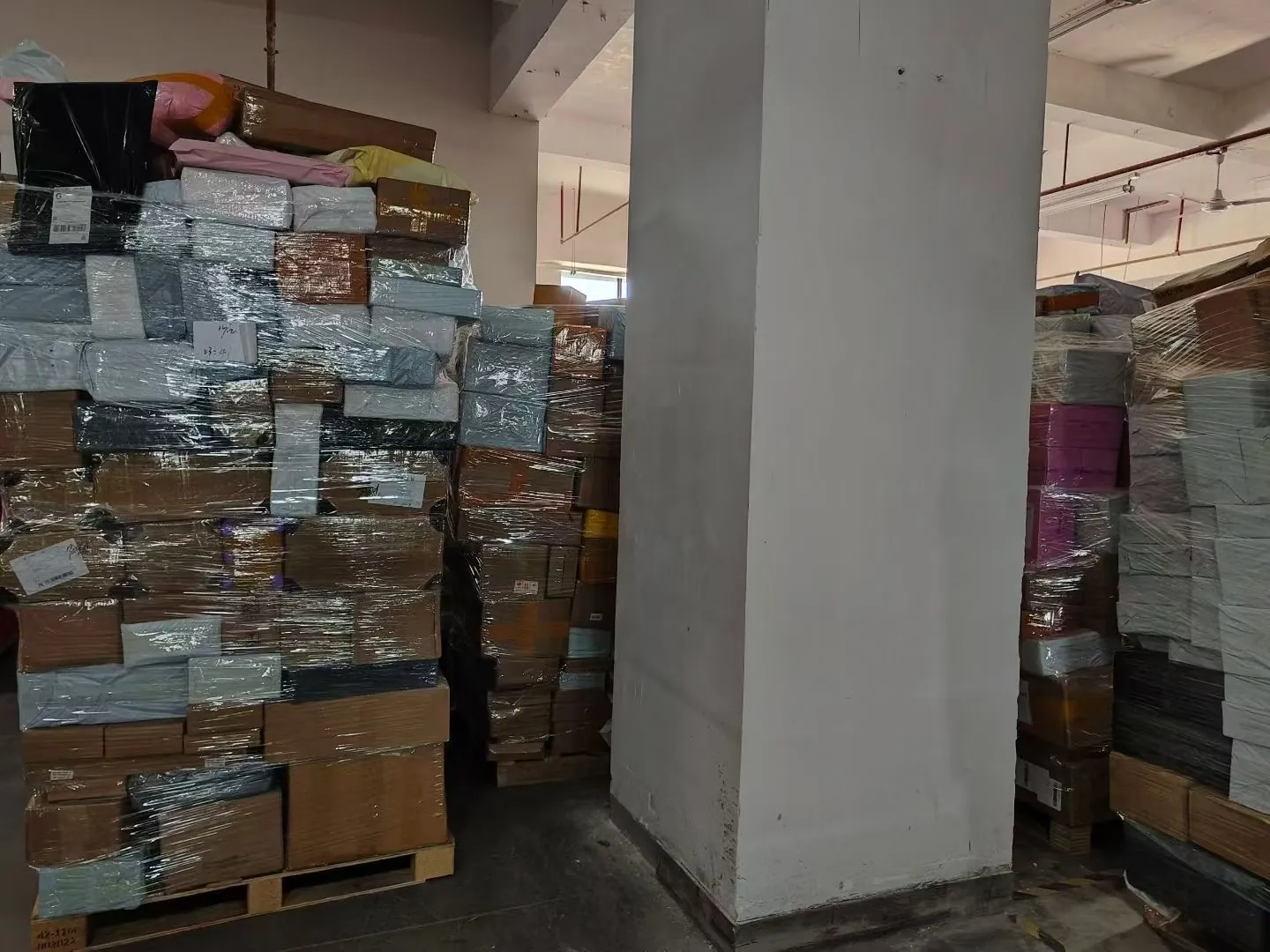 Tall stacks of film-wrapped cartons stored around a concrete pillar in the warehouse
