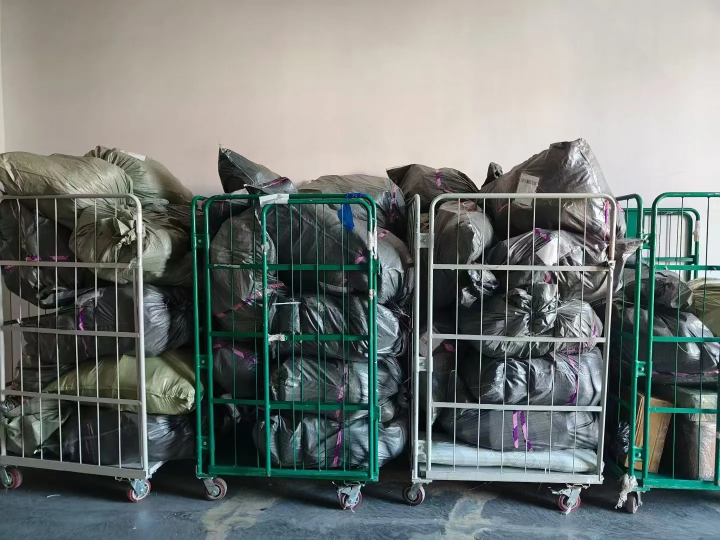 A row of green and silver cage trolleys packed with black sacks of parcels against a warehouse wall