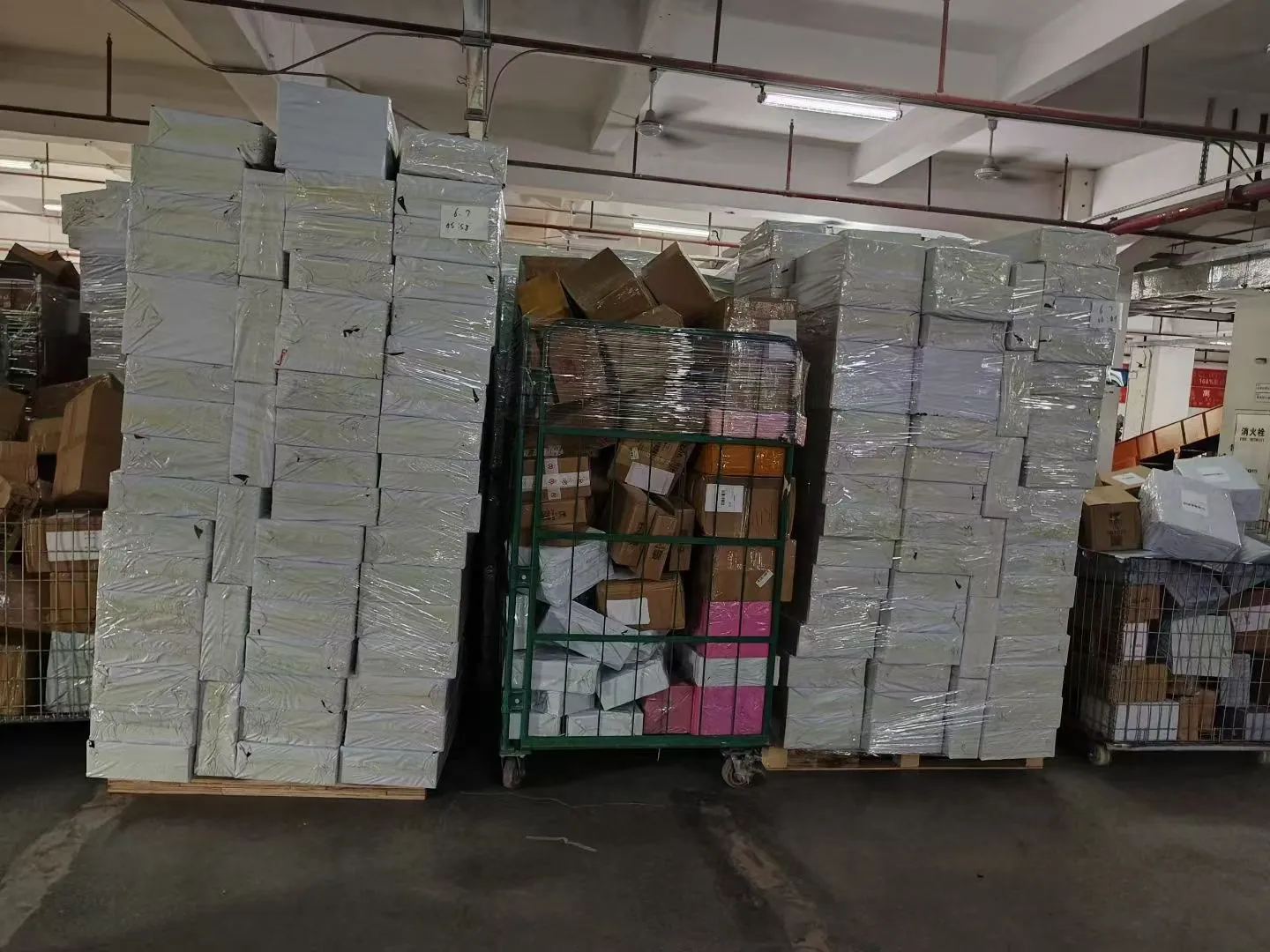 Tall stacks of shrink-wrapped white product boxes stored in the warehouse, with a cage of cartons beside them